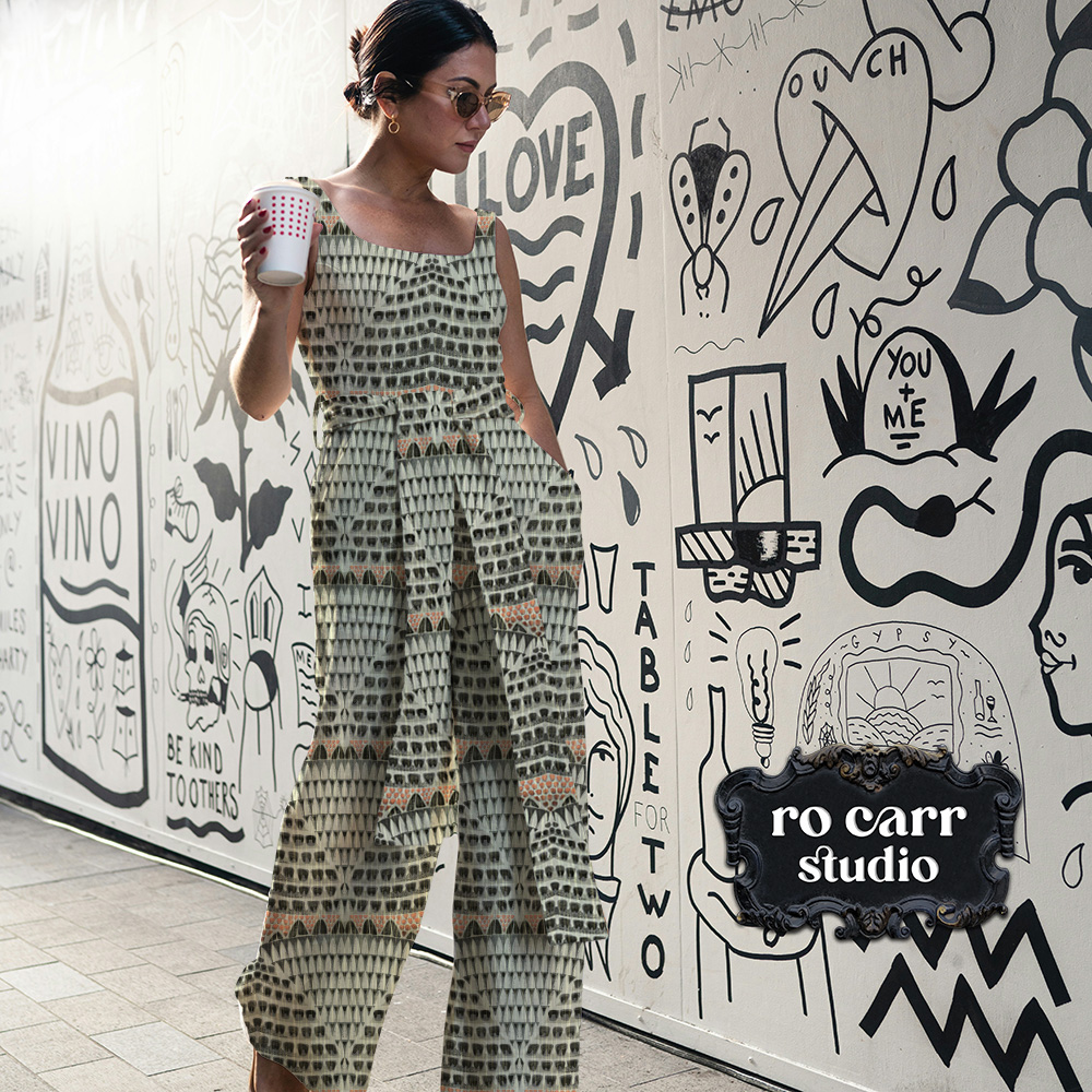Woman walking with a coffee in front of a graffitied wall a sleeveless jump suit made from fabric printed with the Armadillo pattern.