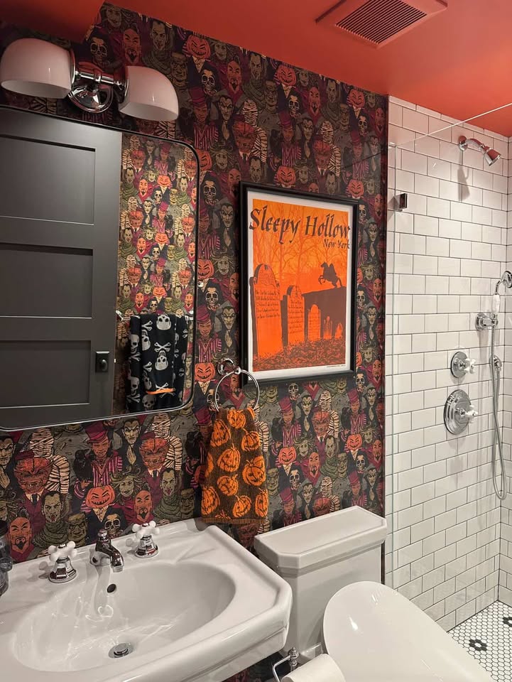 Orange ceiling paired with white subway tile and halloween creatures papering the walls. featured is a Sleepy Hollow framed print and pumpkin and skull printed hand towels.