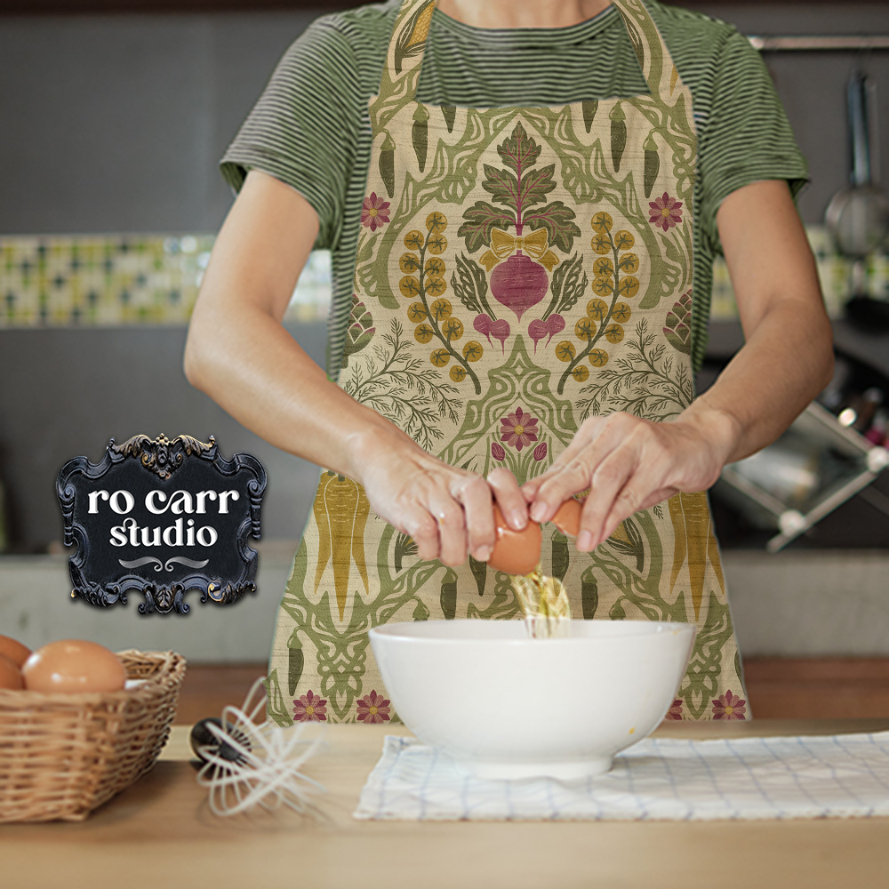 Heirloom & Bloom vegetable pattern printed on a kitchen apron worn by a person cracking eggs over a mixing bowl.