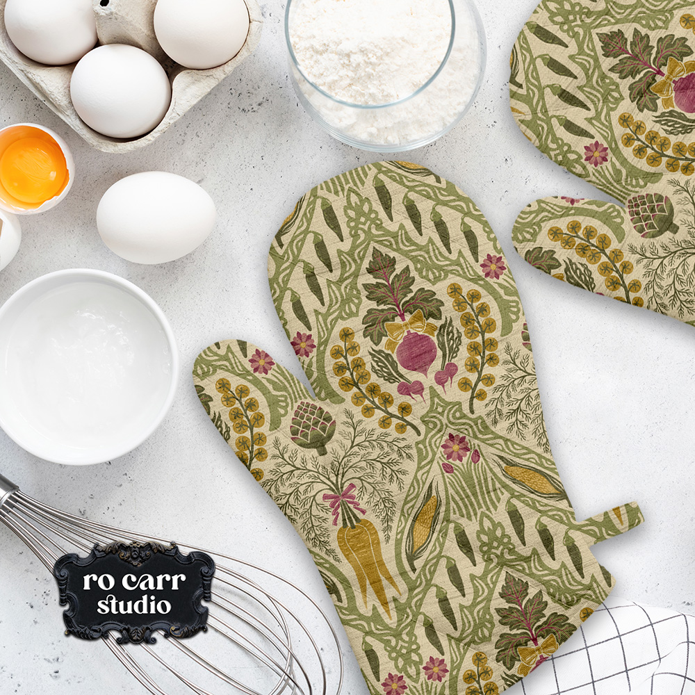 Heirloom & Bloom vegetable pattern printed on oven mitts and kitchen textiles arranged beside eggs, flour, and baking tools.