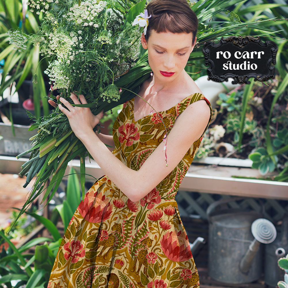 A woman in a full-skirted dress printed with a damask leopard and tropical floral pattern in gold, green, and rose, standing among greenhouse plants.