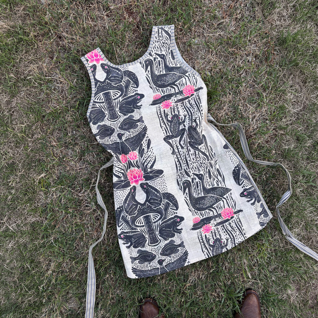 A sleeveless mini dress with a block-print pattern of bullfrogs, lotus blooms, and wading birds, with striped trim detailing.