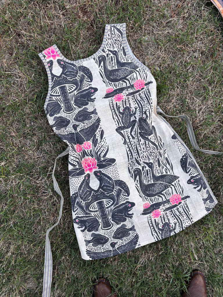 A sleeveless mini dress with a block-print pattern of bullfrogs, lotus blooms, and wading birds, with striped trim detailing.