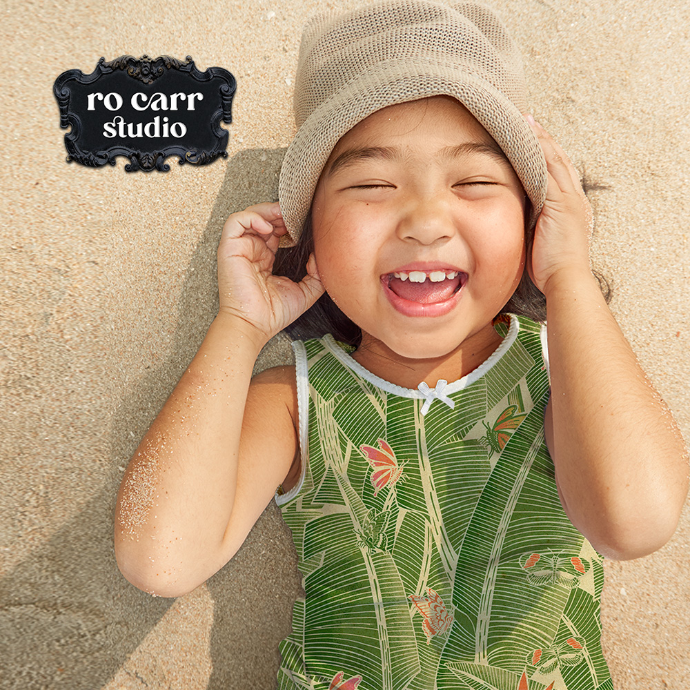 Laughing child in a Goodnight Jungle zip-up garment, lying on sand.